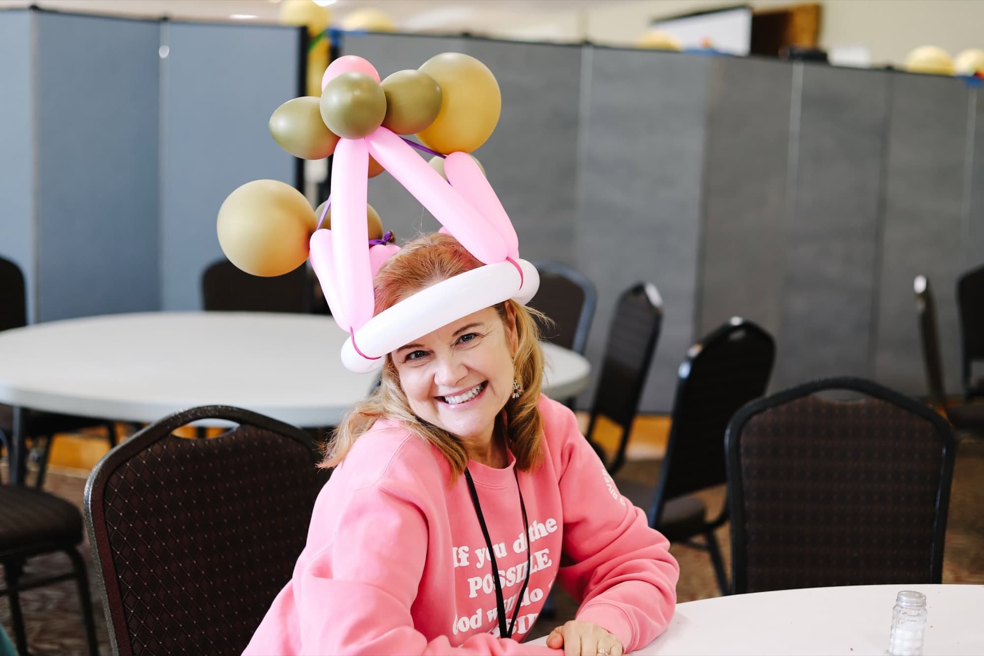 A smiling girl wears a pink balloon hat at a table