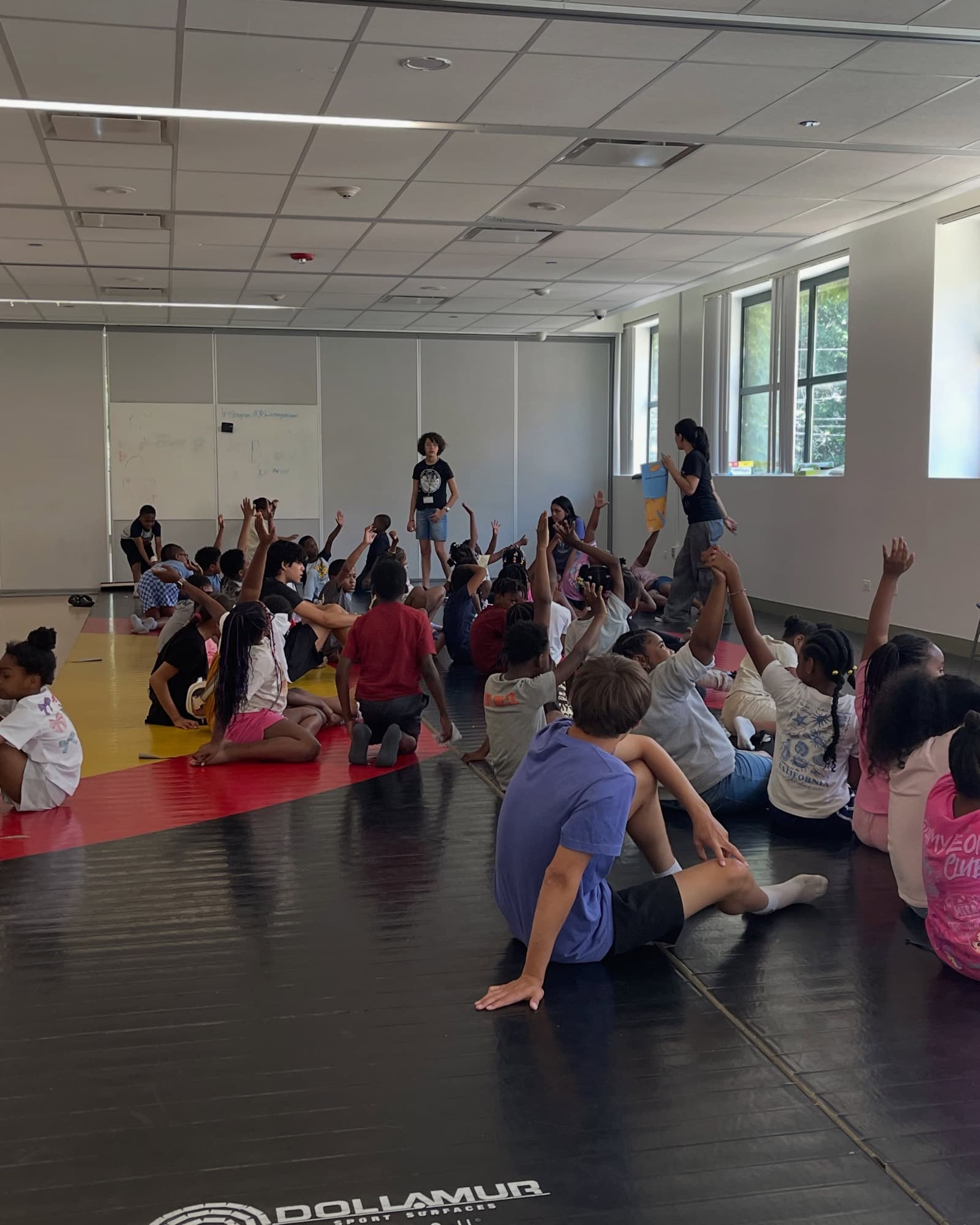 A large group of children sit on the floor of a bright hall while leaders teach