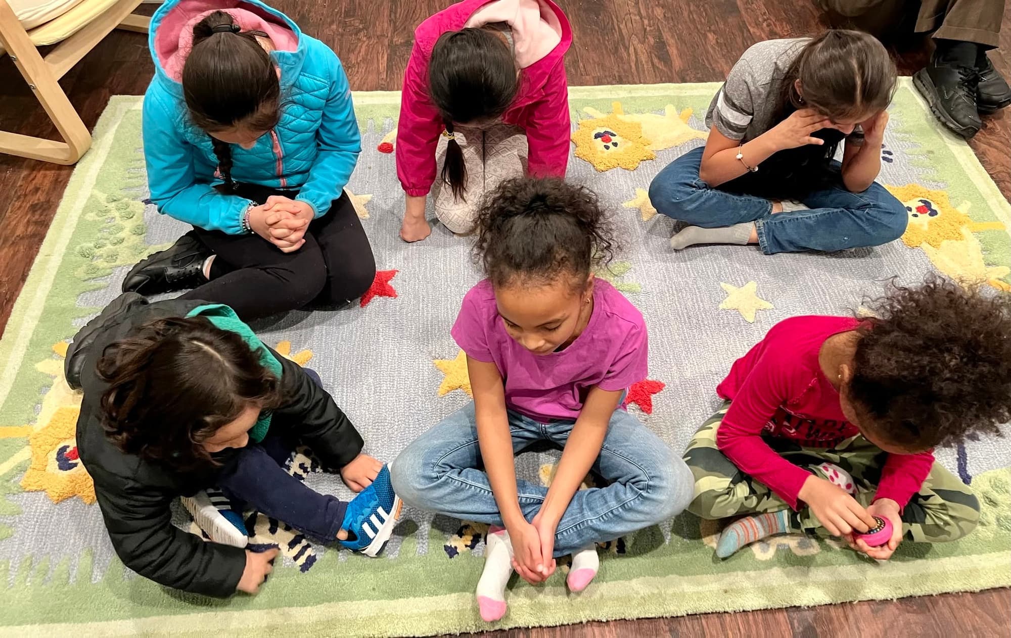 Children sit in a circle on a rug with their heads bowed and hands folded in prayer