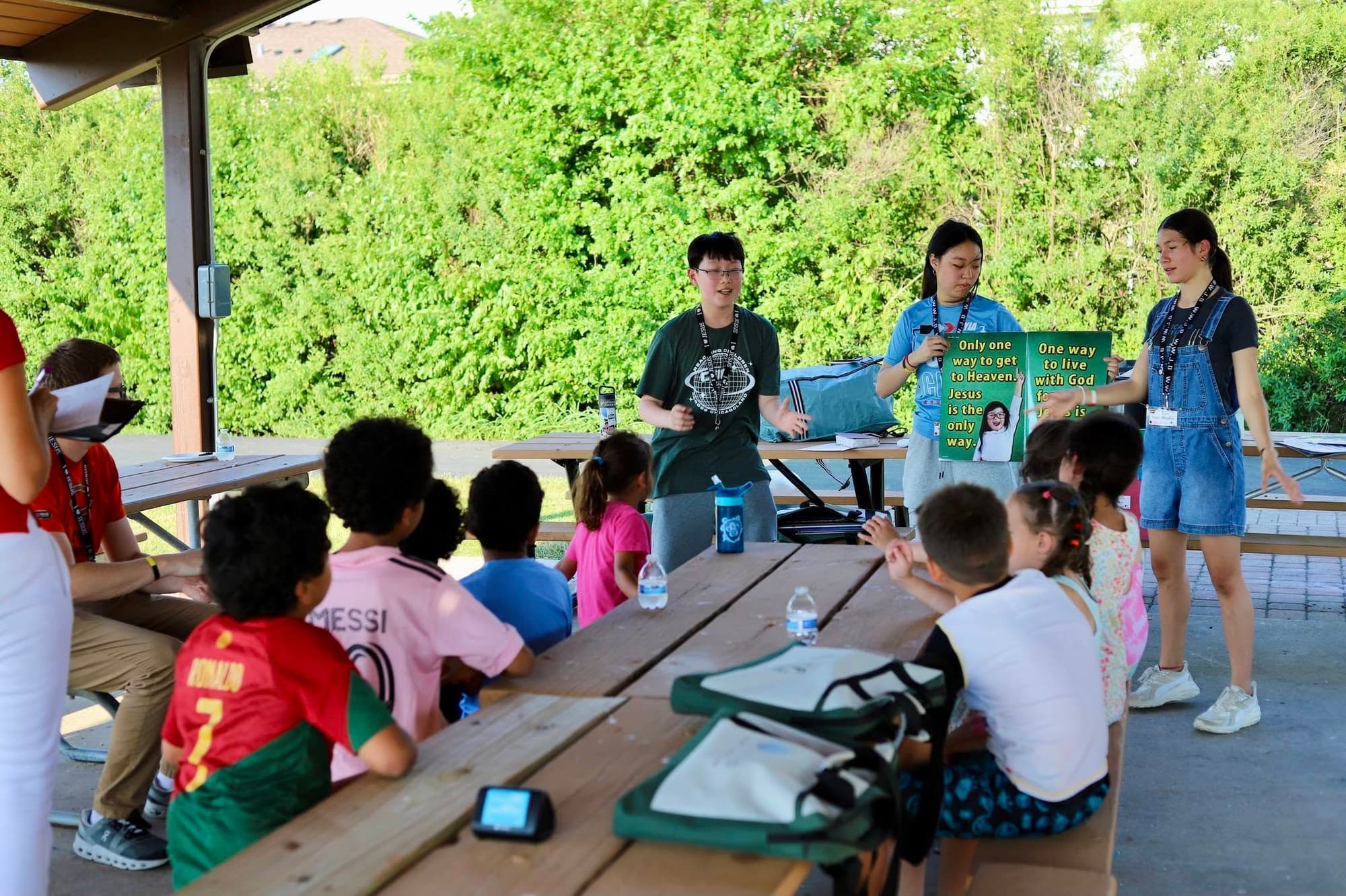Teenage leaders teach children sitting at picnic tables under a park shelter