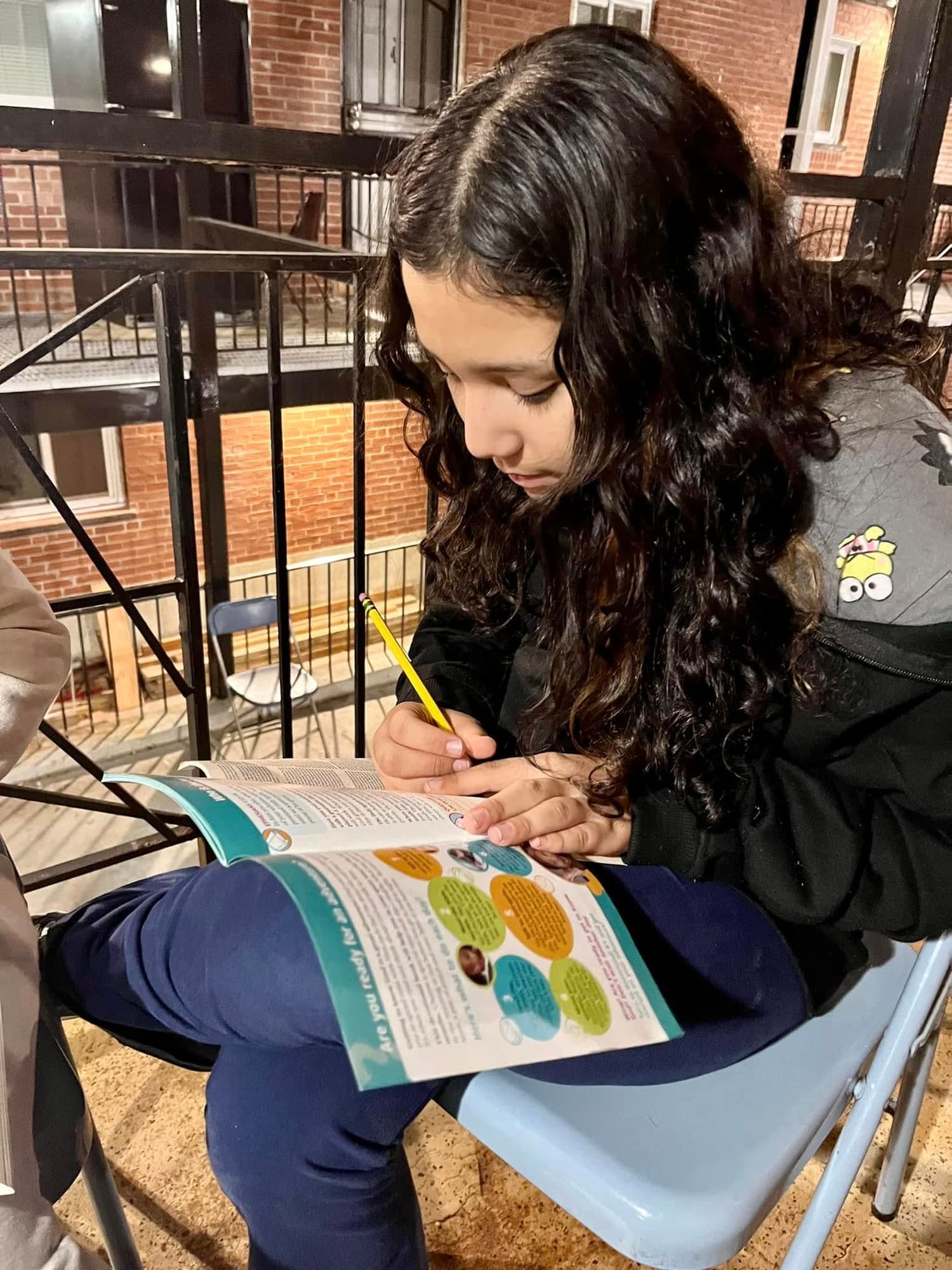 A girl writes in an activity booklet on her lap, sitting outside on a folding chair