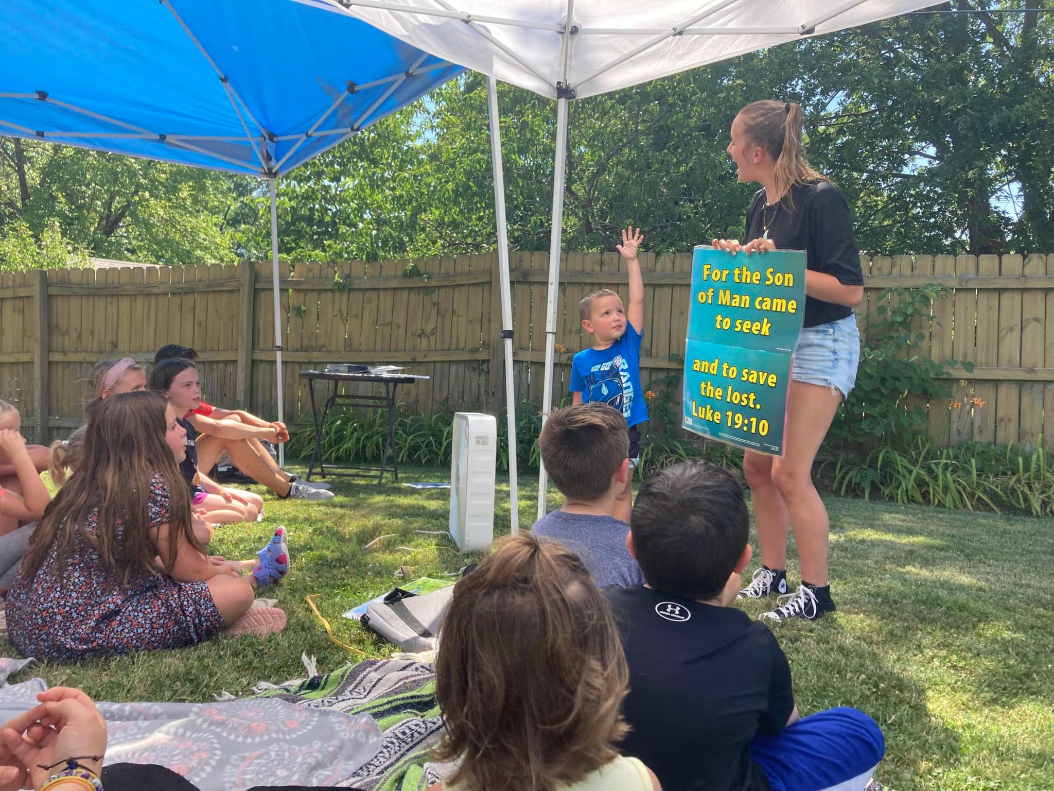 A teacher holds up a memory verse for children sitting on the grass under a tent